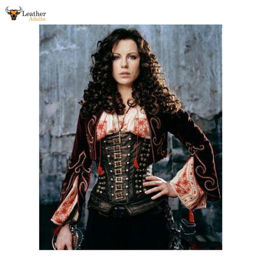 A woman wearing a dark brown and black western-style corset jacket with ornate gold-toned hardware, red trim, and patterned sleeves, posed against a gray stone wall. A Leather Addict logo appears in the top-left corner.