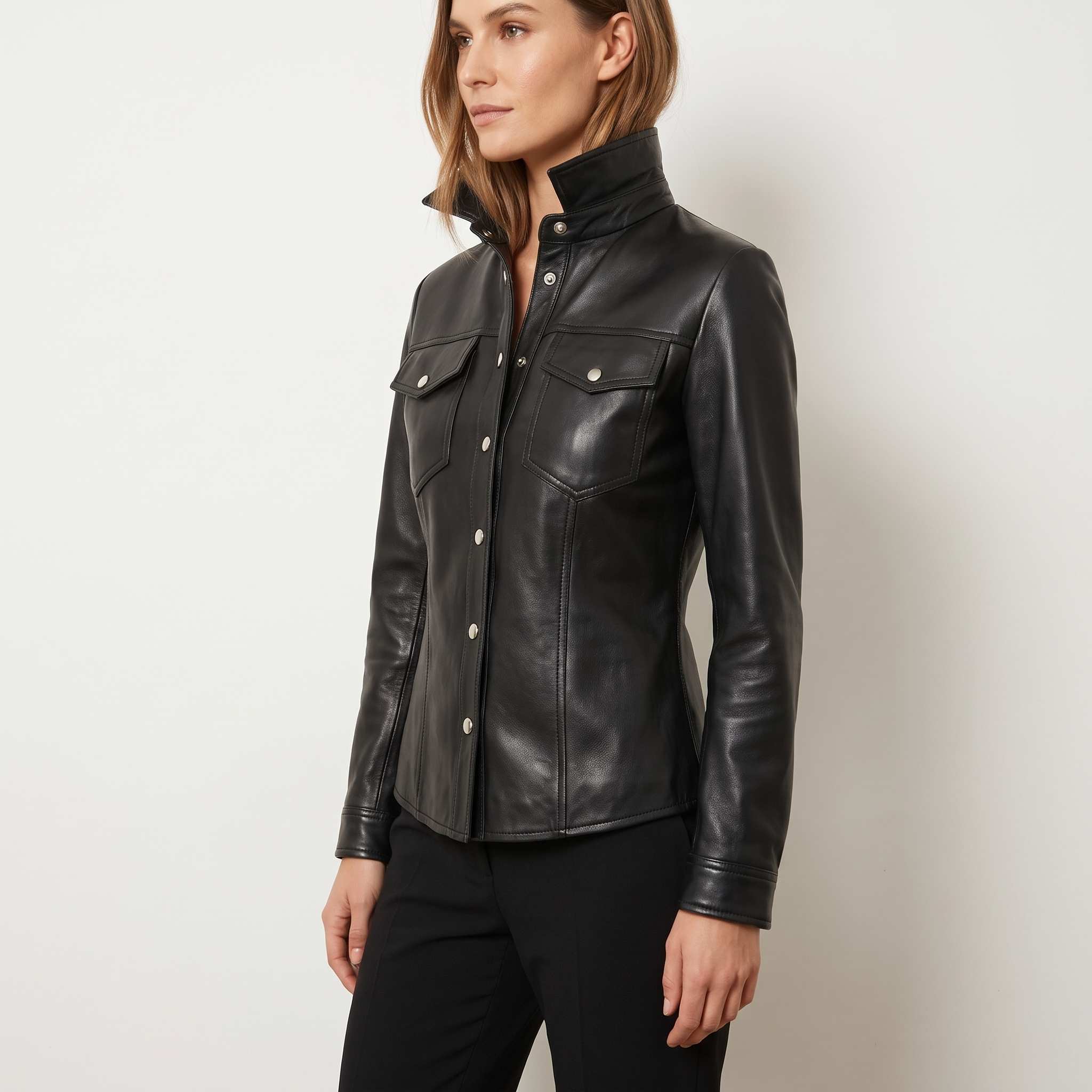 Three quarter side angle of a model posing in a fitted black genuine leather overshirt with a raised funnel collar and silver fasteners.