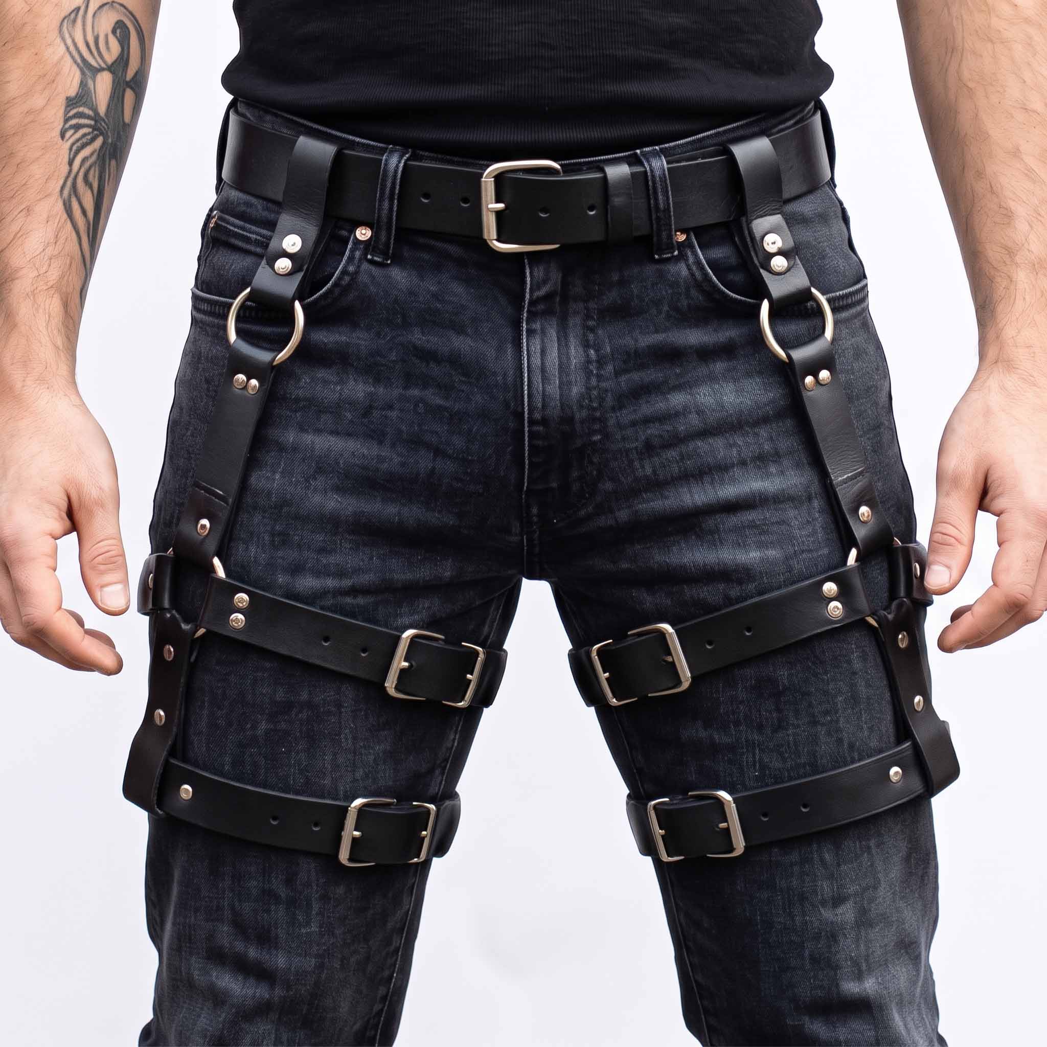 Front view of man wearing handmade black cowhide leather thigh harness with double roller buckle leg straps over black washed denim jeans