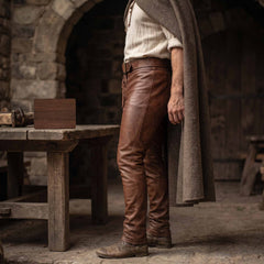 Side profile view of tailored brown leather medieval pants detailing smooth leather grain and tapered leg cut beside a rustic wooden table