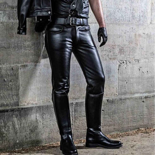 Black faux leather riding pants worn by a model with a black leather jacket, belt, and tall black riding boots, posed against a concrete wall.